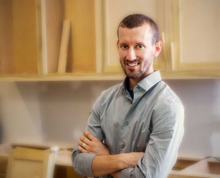 Smiling man with a beard and crossed arms standing in front of unfinished wooden cabinets.