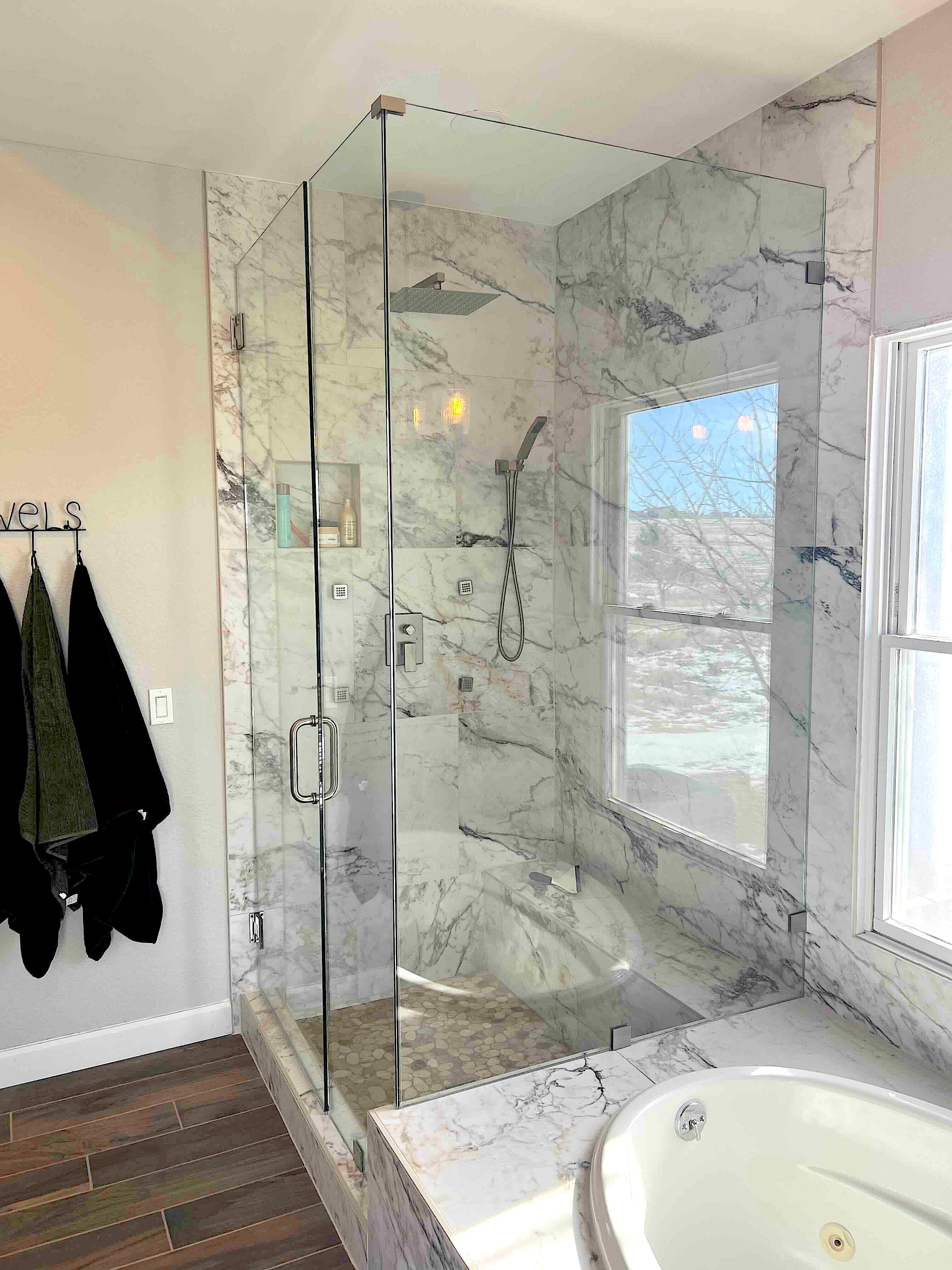 Modern glass walk-in shower with marble walls, pebble floor, and window overlooking snowy hills.